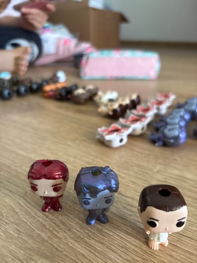 A photograph shows a collection of toys or figurines, some arranged in a line or row on a light-colored wooden surface. At least three figurines are oriented facing up in the frame, these are close to the foreground, while others are arranged to the left and further in the background. The toys or figurines include human figures and animal shapes, possibly dragons. The human figures on the right side have prominent round heads and small bodies, and include details like dark hair, skin tones, and stylized features. Some figures are metallic.
In the background there are boxes and other materials, and a person's hands can be seen. The person is wearing a dark t-shirt and white sleeves.
The exact location in the city of Fulda, Germany cannot be determined from the image content alone. The environment appears to be indoors.