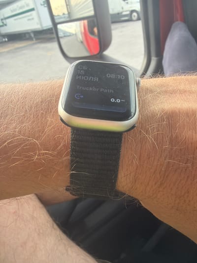 The image shows a close-up of a person's arm wearing an Apple Watch. The watch displays the time as 08:10 and the date as July 18th. It also shows "Trucker Path" with a distance of 0.0 miles. In the background, a semi-truck's side mirror reflects a red semi-truck cab. The environment appears to be outdoors, possibly in a truck stop or parking lot, with other trucks visible in the background, suggesting a transportation hub. The location is the city of Chattanooga, United States of America.
