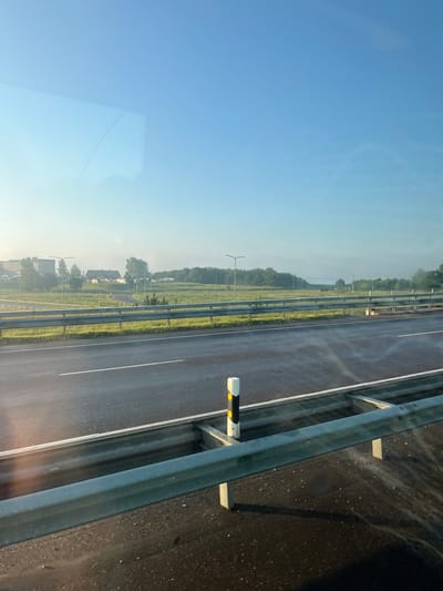 The image shows a highway in Maardu, Estonia, with a clear blue sky and some light fog in the distance. The road is wet, indicating recent rain or dew. A metal guardrail runs along the side of the road, separating it from a grassy median with streetlights. In the background, there are trees, some buildings, and a few small houses. The scene appears to be captured from a moving vehicle, as suggested by the slight blur and the reflection on the window. There are no people or animals visible in the image.