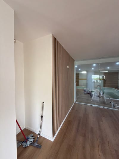 The image captures an interior scene, possibly a hallway or a transitional space, within a building located in Gondomar, Portugal. On the left, a white wall partially obstructs the view, with a silver circular fixture visible near the top corner. The primary focus is a large section of a wall covered in vertical wooden slats, a popular design element. Adjacent to this slatted wall, a light-colored wall continues towards the right. In the foreground, on the wooden floor, rests a cleaning setup comprising a red-handled mop and a silver vacuum cleaner attachment.
Further into the scene, a glass partition reveals another area of the building, possibly an indoor pool or a spa. Inside this glass-enclosed space, a ladder stands with several buckets and painting supplies scattered around it, suggesting ongoing renovation or maintenance work. The flooring in this area appears to be the same wooden material as in the foreground. The lighting in the scene is a mix of overhead lights and ambient light reflecting off surfaces. A silhouette of a person can be seen through the glass, facing away from the camera, and another shadow is cast on the wall within the pool area. The environment appears to be a modern indoor setting, possibly residential or commercial, with a focus on clean lines and contemporary design.
