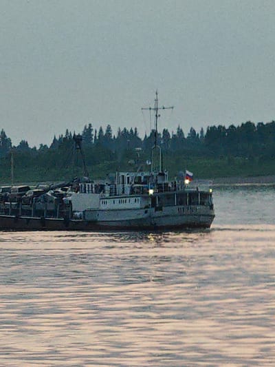 The image shows a cargo ship navigating a body of water, likely the Vop River, in Yartsevo, Russia. The sky is overcast, suggesting either early morning or late evening. The ship, a substantial vessel with a light-colored superstructure, is laden with cargo, which appears to be industrial materials or scrap metal, piled high on its deck. A Russian flag is visible on the stern of the ship. The water is rippled, reflecting the muted light. In the background, a dense line of evergreen trees forms the shoreline. The overall impression is one of industrial activity in a natural, somewhat subdued setting.