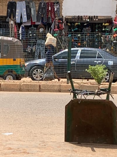 In Bukuru, Nigeria, a street scene unfolds with a focus on commerce and transportation. A man, carrying a large bundle of goods on his head, walks beside a parked dark sedan. To his left, a three-wheeled vehicle, common in local transportation, is partially visible. The background reveals a bustling market stall overflowing with clothing, particularly jeans, and an adjacent shoe shop displaying various footwear. The entire scene is framed by a wire fence, a common feature in many Nigerian street markets, suggesting a lively, open-air retail environment.