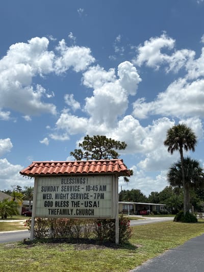 This image shows a church sign on a sunny day in Bradenton, Florida. The sign is white with black lettering and has a red tiled roof. It reads "BLESSINGS! SUNDAY SERVICE - 10:45 AM WED. NIGHT SERVICE - 7 PM GOD BLESS THE-USA! THEFAMILY.CHURCH". The sign is located in front of a building that appears to be a mobile home, with palm trees and other vegetation visible in the background. The sky is blue with fluffy white clouds.