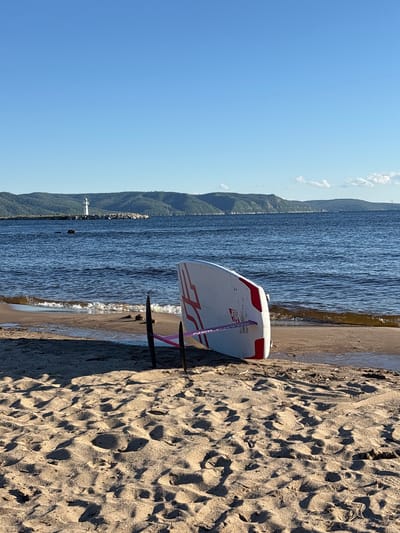 An outdoor scene depicts a sandy beach with a lake or river in the background. The foreground is dominated by sand, with ripples and shadows indicating a sunny day. A white and red windsurfing board is propped up on its side in the sand, angled towards the water. The board has a pink fin attached. Small waves lap at the shore. In the middle ground, the water stretches out towards a distant shoreline with green, tree-covered hills. A white lighthouse with a stone base sits on a jetty in the water, extending out from the far shore. The sky is a clear, bright blue. The location is the city of Volzhskiy Utes, Russia.
