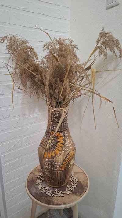 This image shows a close-up of a decorative ceramic vase filled with dried reeds. The vase is an ornate, hand-painted object with a warm, earthy color palette, featuring a prominent sun-like motif in yellow and orange. The reeds are light brown and feathery, creating a soft, natural texture against the detailed pattern of the vase. The vase itself rests on a wooden stand, which has a circular base and a decorative wooden overlay of a floral garland.
The background consists of a white textured wall that resembles brickwork. The ceiling is also white and shows a simple light fixture. The lighting appears to be indoor, ambient light, suggesting the scene is set within a home or decorative display area. There is no indication of time of day or weather. The overall impression is one of rustic, natural decor. The image is taken from a slightly low angle, looking up towards the vase. No people are visible in the image, and there is no visible text. The context provided is Cherkessk, Russia, suggesting this is a decorative item displayed in that location.