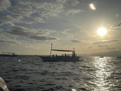 The scene depicts an open body of water off Pengastulan, Indonesia, during either sunrise or sunset. The sun, a bright circular orb, is positioned in the upper right quadrant, emitting strong light that causes significant glare and a bright reflection path across the dark, choppy water. A distinct, crescent-shaped flare or reflection is visible in the upper center-left sky. The sky is partially cloudy with scattered stratocumulus clouds.
In the mid-ground, a light-colored traditional Indonesian boat, equipped with an overhead canopy supported by poles and displaying red and white flags on its forward pole, is central. Approximately 4-6 individuals are visible within this boat as dark silhouettes under the canopy. A lifebuoy is attached to the port side of this vessel. To the left of the central boat and further back, a second similar traditional boat is visible, also with a canopy and indistinct occupants. Multiple other smaller boat silhouettes are scattered across the distant horizon.
The far background on the right features a low-lying mountain range or series of hills silhouetted against the bright horizon. The extreme bottom-left foreground shows a dark, curved object, consistent with a section of the boat from which the image was captured.