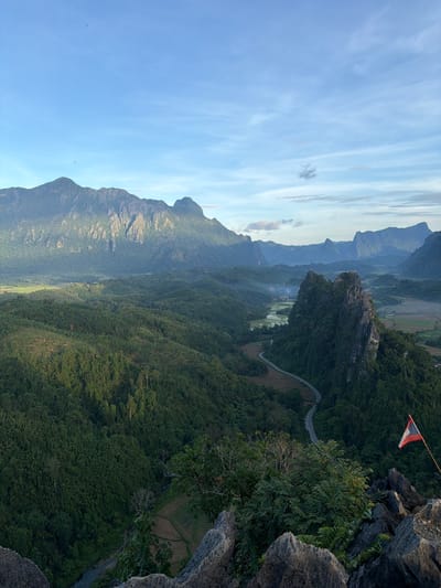 This is a landscape photograph taken from a high vantage point, overlooking a lush, verdant valley. The scene is dominated by rolling hills covered in dense green forest, with jagged, rocky outcrops interspersed throughout. In the distance, a winding road snakes its way through the valley, bordered by rice paddies. The sky is a clear blue, with a few wispy clouds scattered across it. A small flag, appearing to be the flag of Laos, is visible in the foreground, planted on a rocky ledge. The overall impression is one of natural beauty and tranquility. The image does not contain any people or specific actions. The location is described as being in Naka, Laos.