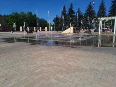 The image depicts a sunny day in a city square in Otradny, Russia, featuring a modern fountain area. The foreground is dominated by a patterned light-colored brick pavement, wet from the fountain's spray. Multiple jets of water erupt from the pavement in the midground, creating a dynamic visual. Behind the fountain, benches are visible, and a bicycle is parked. Further back, evergreen trees line the square, with a low, angled, light-colored building with yellow accents to their right. On the far right, a white pergola structure is present, under which two young girls are being pushed on a swing. Two women are standing nearby. In the distance, more trees and buildings can be seen under a clear blue sky. The scene suggests a public recreational space in use.