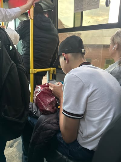 The image depicts the interior of a bus in Smolenshchina, Russia. Several individuals are visible, some of whom are seated and others who appear to be standing. The scene is crowded, with passengers occupying various seats and potentially the aisle. The time of day appears to be daylight, suggested by the lighting through the bus windows, though the specific time is not discernible. The weather conditions outside are unclear from the interior view.

One prominent subject is a young person wearing a black baseball cap and a white t-shirt with red and black stripes on the collar. This individual is looking down, possibly at a phone or in conversation with someone out of frame. They are wearing earbuds. Another person, partially visible, has blonde hair and is also wearing earbuds. In the foreground, a person in a white shirt is holding onto a yellow pole, a common fixture in public transport.

Several objects are present, including a dark-colored backpack or bag on the floor, a maroon plastic bag held by a person, and what appears to be a mobile phone. The bus itself features yellow poles for passenger support and dark-colored seating. A document or notice is visible on a panel near the window.

The overall atmosphere seems to be one of everyday commuting. There are no overt indicators of a specific event or strong emotions, suggesting a typical journey. The presence of multiple individuals and the bus interior confirm the setting as a public transportation vehicle.