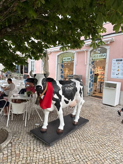The image captures an outdoor scene in Sintra, Portugal. An artificial, black-and-white, cow statue bearing the name "ALE-HOP" stands near an "ALE-HOP" cafe with a pink facade. The statue sports a red cloth and a bell. People sit at white tables and chairs next to the statue, appearing to be patrons of the café. The foreground features a tree with dense green foliage. The ground is paved with small, light-colored stones. The sky is not visible.
