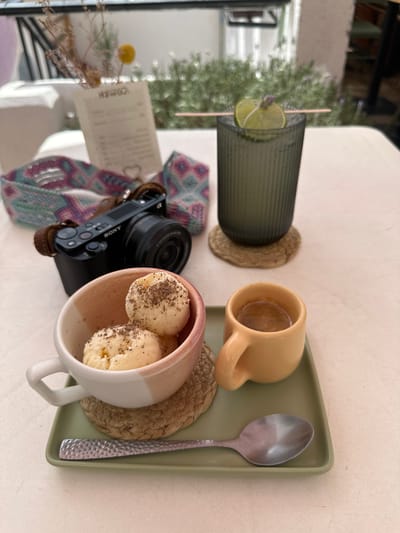 A table in an outdoor cafe in Zacatecas City, Mexico, displays a spread of food and drink. A Sony camera with a leather strap rests on the table near a decorative woven band. In the foreground, two scoops of ice cream are served in a pink-rimmed white cup atop a woven coaster. Beside the ice cream is a small yellow mug containing a dark liquid, likely coffee. A silver spoon lies on a green tray in front of the cups. In the background, a tall glass with a lime and lavender garnish contains a beverage, and a menu is propped up behind the camera. The setting suggests a relaxed outdoor dining experience.
