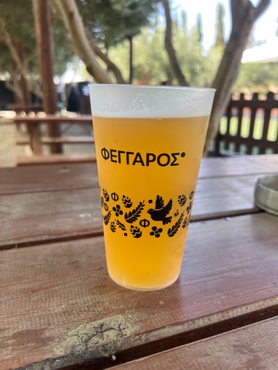 A close-up shot shows a pint glass filled with a golden-hued beverage, topped with a frothy white head, resting on a weathered wooden table. The glass is adorned with black graphics, featuring the word "ΦΕΓΓΑΡΟΣ" in Greek lettering at the top, followed by a decorative pattern of leaves, flowers, and a bird. The background, out of focus, reveals an outdoor setting with trees, foliage, and what appears to be a wooden fence or railing. The overall impression is that of a relaxed, possibly outdoor dining or drinking establishment, likely in a rural or semi-rural location. The specific location in Analiontas, Cyprus is inferred from the context provided, suggesting a natural or agricultural environment.