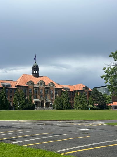 The image captures a frontal view of a large, historic brick building with a red tile roof, likely an institutional or educational facility, situated in Sainte-Anne-de-Bellevue, Canada. The building features a prominent central tower topped with a cupola and a flag, which appears to be dark blue with white lettering. The architecture includes arched windows, decorative stone accents, and a grand entrance with a set of stairs.
The building is set back from a manicured green lawn, which is a significant portion of the foreground. In front of the lawn, there is a paved area, possibly a parking lot or driveway, showing signs of recent rain with puddles and wet patches. Yellow and white lines delineate parking spaces.
To the right of the main building, a modern glass-fronted structure is visible, suggesting a contrast between old and new architecture within the same complex or nearby. Mature trees, primarily evergreens, are strategically planted around the building and on the lawn, adding to the established feel of the landscape. The sky above is overcast with dark, heavy clouds, indicating potentially inclement weather. There are no people visible in the image.