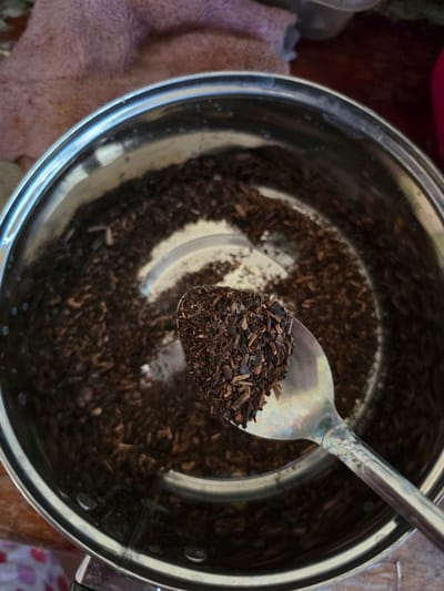 An overhead medium shot shows a stainless steel pot filled with dark, granular tea leaves. A silver spoon rests at an angle, holding a scoop of the same tea leaves, positioned over the pot. The rim of the pot reflects light, and a few small water droplets are visible on the inner surface. A textured pink cloth is partially visible in the upper left corner of the image, and a wooden surface is beneath the pot. The image appears to be taken indoors. There is no information available about the location in the city of Pintomakmur, Indonesia.