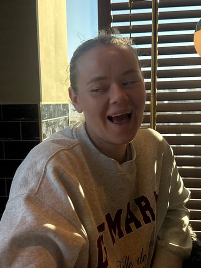 A close-up, indoor photograph taken in Mysiadło, Poland, depicts an adult female subject from the chest up, facing forward. She has an open-mouthed expression, characteristic of laughter or a shout, with her eyes partially squinted. Her light-colored hair is pulled back, with wisps visible around her head. She wears a light grey crewneck sweatshirt featuring dark red lettering, partially reading "MAR" and "Ville de" below it. Direct sunlight, entering from the right, illuminates her right side and casts shadows on the left side of her face and torso. The background on the left consists of a pale yellow-beige vertical surface above a section of dark, possibly black, rectangular tiles. On the right, horizontal wooden blinds are visible, with bright sunlight filtering through them, and a portion of a spherical, opaque white light fixture is present in the upper right quadrant.