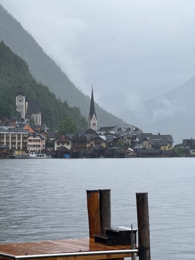 The image depicts a view of Hallstatt, Austria, on a rainy day. In the foreground, a wooden dock with metal railings and wooden posts is visible, with water rippling in front of it. The middle ground features the picturesque town of Hallstatt, with its colorful buildings, including a church with a tall spire. The buildings are situated along a waterfront, with smaller wooden structures and boats docked along the shore. Behind the town, a lush green mountain rises steeply, partially obscured by mist and rain. The overall atmosphere is moody and serene, typical of a cloudy, wet day in this historic Austrian village. There are no people visible in the scene.