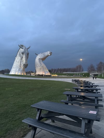 The image depicts an outdoor scene at The Helix Park in Falkirk, United Kingdom, featuring The Kelpies sculptures at dusk or dawn under an overcast, dark grey sky. In the mid-ground, two massive, stylized horse head sculptures, constructed from reflective metallic panels, stand illuminated from below. The left sculpture faces generally forward, while the right sculpture's head is angled upwards and to the right. Both sculptures are situated on circular bases surrounded by paved areas and manicured green grass.
In the foreground, a row of approximately eight dark-colored, unoccupied wooden picnic tables with attached benches extends from the lower right towards the mid-ground, set on a grey paved surface. One table in the immediate foreground displays a white sticker with a red prohibition symbol. The ground transitions to a well-maintained green grass lawn to the left.
In the background, beyond the sculptures, a paved pathway is visible. Four human figures are present: two indistinct individuals near the base of the left sculpture, and two individuals walking away from the camera along the pathway in the far right mid-ground. A tall streetlamp with a bright light is visible to the right of the sculptures. A line of bare deciduous trees runs horizontally across the distant background, with power lines and other urban infrastructure faintly visible on the far left. The overall lighting is low, emphasizing the illuminated sculptures against the dark, featureless sky.
