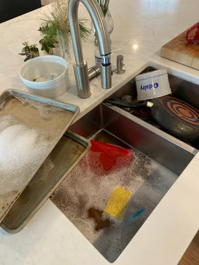 A stainless steel kitchen sink with two basins. The left basin contains a baking sheet filled with soapy water, while the right basin has a red silicone spatula and a yellow sponge in soapy water. A white plastic container with the word "Dairy" printed on it and a black frying pan are in the right basin. There is a silver faucet in the middle, along with a white porcelain bowl and a silver soap dispenser. The counter top is white marble. This is located in Rye, New York, United States, at latitude 40.952 and longitude -73.708. The scene appears to be in the middle of dish washing.