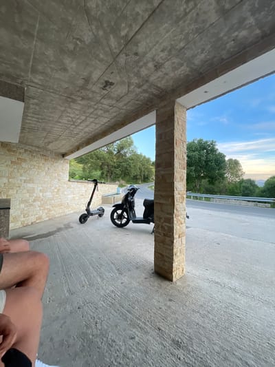 The image depicts an outdoor scene, likely a covered area with a concrete floor and a stone-clad wall on the left. Above, a textured concrete ceiling is visible. Two vehicles are parked on the concrete: an electric scooter on the left and a black scooter on the right. In the background, a paved road curves through a landscape of green trees under a bright blue sky with some clouds. The location is identified as Valkosel, Bulgaria. A person's legs and lower torso are visible in the foreground on the left, suggesting they are seated or standing, partially obscuring the view. The overall impression is that of a stop along a scenic route.