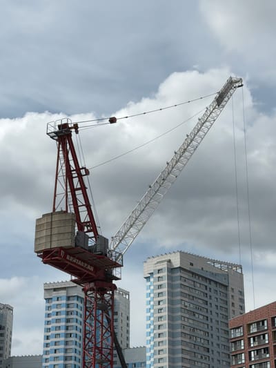 This media file is a photograph capturing a construction site with multiple buildings under development. The primary focus is a large white lattice boom crane that extends diagonally across the frame. Attached to the main boom is a red crane jib, which is counterbalanced with concrete weights. The jib has a section with visible text reading "REATAKPAN". A person is visible standing on the top of the white crane's boom.
In the background, several multi-story buildings are visible. One building on the right is made of reddish-brown brick with numerous windows. Adjacent to it is a taller, modern building with a gray and blue facade, also featuring many windows arranged in a grid pattern. Another building with a blue and white striped pattern is also present. The sky is overcast with dramatic clouds, suggesting a cloudy day. The lighting is diffused, indicating it is likely daytime, but the exact time cannot be determined. The scene appears to be a large-scale construction project in an urban or suburban setting. There are no explicit emotional cues, and the overall atmosphere is one of ongoing construction and development.