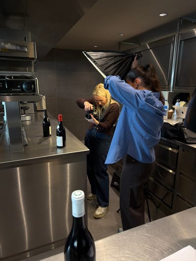 The scene depicts a product photography session in a modern, industrial-style kitchen in Marche-lez-Écaussinnes, Belgium. Two females are engaged in photographing wine bottles. A blonde female, positioned slightly to the left, is holding a black DSLR camera with both hands, aiming it downwards towards the counter. She wears a brown long-sleeved top and dark pants. To her right, a second female with dark hair tied in a high ponytail holds a black rectangular softbox light modifier, directing light towards the subjects. She is dressed in a blue and white checkered long-sleeved shirt and dark pants.

On the brushed stainless steel counter in the mid-ground, three dark glass wine bottles are arranged, two with dark foil caps and one with a red foil cap and a white label. In the immediate foreground, a fourth dark glass wine bottle with a white foil cap is partially visible on a lower stainless steel surface. The kitchen features extensive stainless steel countertops, cabinets, and professional appliances, including a large oven-like unit and another machine with a prominent dial. The walls are dark gray, and recessed lighting is visible in the ceiling. A MacBook laptop and a water bottle are present on a distant counter to the right. A black stool or low tripod is partially visible near the feet of the female holding the softbox.