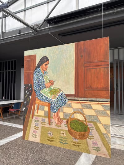 A large, rectangular canvas painting is displayed vertically outdoors in the city of Drama, Greece. The painting is suspended by thin lines from a dark, horizontal track system, which is integrated into a metallic truss structure supporting a translucent roof overhead. The ground beneath is a dark gray speckled aggregate pavement, featuring a prominent, broad light-colored diagonal stripe.
The painting depicts an elderly woman seated in a domestic setting. She wears a dark patterned headscarf and a long-sleeved, blue, green, and white patterned dress, and is barefoot. She sits on a simple wooden chair with a woven seat, leaning forward as she uses a small knife in her right hand to prepare green leafy vegetables held in her left hand. A quantity of these vegetables rests on a patterned green and white apron or cloth covering her lap. To the woman's right, on a floral patterned rug, a woven basket filled with green vegetables is present. The rug features pastel-colored floral motifs (pink, blue, white) on a light green background. The floor in the painting is composed of light blue-grey and light yellow-beige checkered tiles, delineated by a narrow red and black striped border. The background within the artwork includes a light blue-green wall to the left and a reddish-brown wooden door with paneling and a visible keyhole on the right. A dark fabric with white animal silhouettes is partially visible to the left of the woman's chair. A signature is present in the lower right corner of the canvas.
In the actual environment surrounding the painting, to the left, a smooth light grey wall and a dark, vertically barred window are visible, alongside portions of a white table and two chairs (one blue, one red). To the right of the painting, a light marble step leads to a partially open, grey-framed glass door, offering a glimpse into an interior space.