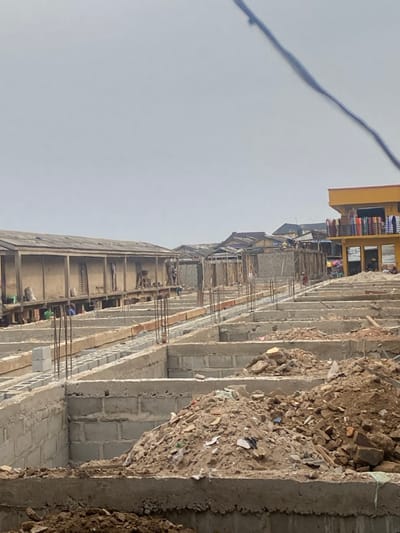 An unfinished building stands under a bright sky, constructed with layers of cement blocks and reinforced with rebar. Construction debris piles up along the side, adding to the unkempt atmosphere. In the background, a yellow building with colorful items displayed on its balconies suggests nearby commercial activity. The entire scene presents an active construction site.