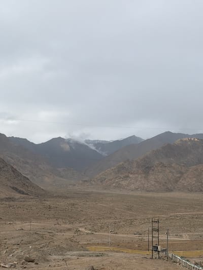 This is a landscape photograph taken in Chumkar, India, under a cloudy sky. The image captures a vast, arid valley with rugged, barren mountains in the background. The mountains are layered in shades of brown and grey, with some peaks appearing snow-capped. In the foreground, the ground is dry and dusty, scattered with rocks and sparse vegetation. A yellow-walled enclosure is visible in the lower right portion of the frame, next to a metal utility pole with electrical equipment. Power lines are strung across the sky, bisecting the view of the mountains. There are no people or animals visible in the image. The overall scene depicts a remote, undeveloped, and harsh environment.