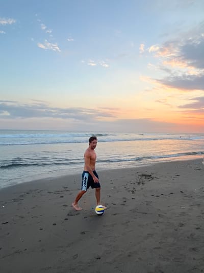 A shirtless man wearing dark blue and white patterned swim trunks is walking on a sandy beach, kicking a volleyball. The sun is setting, casting a warm orange and pink glow across the sky and reflecting on the ocean waves. The beach is dark gray sand with footprints. In the distance, several people can be seen in the water. The scene is located in North Kuta, Indonesia.