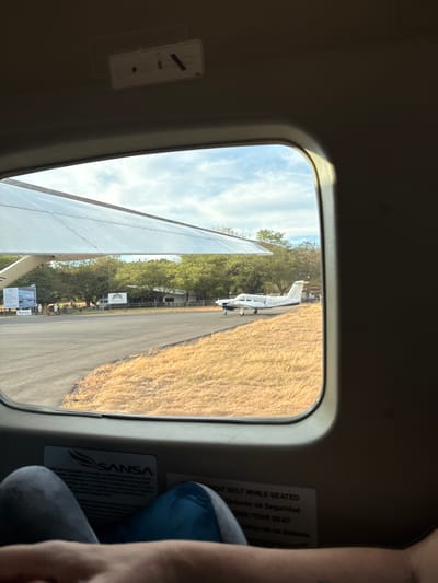 The image shows a view from inside a small airplane through a window. The wing of the plane is visible along with the runway and a small white plane parked alongside it. The runway is bordered by dry grass and trees. The window frame and interior of the plane are visible. A seatbelt instruction card with the word "SANSA" is affixed to the plane's interior. Part of a person's arm and leg is visible on the right side of the frame. The OCR text shows the registration of the white plane along the runway as "TI-AN". The seat belt instructions are in both English and Spanish. Villarreal is in Costa Rica, it is likely that this image was taken somewhere near Villarreal.