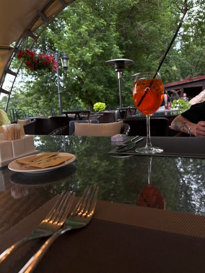 The image depicts an outdoor seating area, likely at a cafe or restaurant, in Izmailovo, Russia. A table with a reflective surface is in the foreground, set with two forks, a glass containing an orange beverage with ice and a straw, and a plate with two small packets and a dispenser of toothpicks. A cigarette pack and a knife are also visible on the table. In the background, there are tables and chairs, some with cushions, suggesting a seating arrangement for patrons. Lush green trees and foliage dominate the background, indicating a natural or park-like setting. A lamppost with a hanging basket of red flowers and a gas heater are also present. The overall atmosphere suggests a relaxed outdoor dining experience.