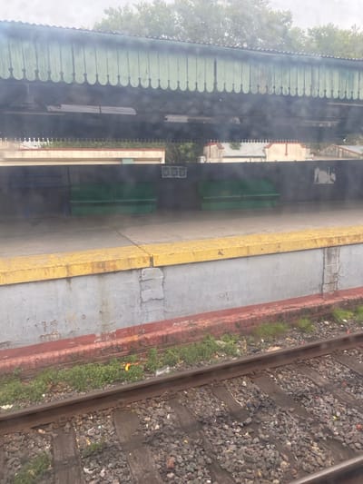 The image displays a train station platform and tracks within the city of Partido de Lanús, Argentina, viewed through a wet or smudged window, creating a hazy effect. In the foreground, two parallel train tracks with dark gray ballast and visible sleepers run horizontally. Green vegetation, along with scattered small debris including a yellow object and white plastic, grows between the tracks and the platform edge.
Above the tracks, a concrete platform wall, predominantly light gray and weathered, features a dark red horizontal strip along its base. The top of the platform is defined by a yellow concrete safety strip, showing signs of wear and dirt. The platform surface is concrete, appearing damp or stained, with a central longitudinal seam. On the platform, against a dark gray wall, two sets of backless, three-seat green benches are positioned. Faint markings are visible on the dark wall behind the benches.
Above the platform, a light green, scalloped canopy extends overhead, covering part of the platform. Beyond the platform and canopy, various buildings with light-colored walls and some red-tiled roofs are visible, partially obscured by a dense line of green trees or foliage. The sky is uniformly overcast. No people are present in the scene.