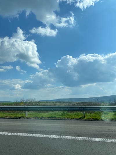 The image shows a view from a vehicle traveling on a road, likely a highway, in or near Zlatina, Bulgaria. The right side of the frame is dominated by the road surface, with white dashed lines marking the edge of the lane. Beyond the road is a grassy shoulder, a guardrail, and a landscape with trees and what appears to be a distant ridge or mountain range. The left side of the frame displays a bright blue sky with puffy white clouds, which occupy a significant portion of the image. The overall composition indicates a sunny day with clear visibility. There are no people visible in the image.