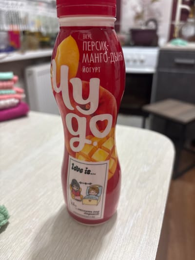 This is a horizontal, eye-level, medium shot of a red plastic bottle of yogurt with a white lid. The bottle is lying on its side on a light-colored, wooden surface, possibly a table. The background is blurred, showing what appears to be a kitchen or dining area with some appliances and cabinets.
The bottle has a label with text and graphics. The prominent text on the label is in Russian and includes "ВКУС ПЕРСИК-МАНГО-ДЫНЯ" (Taste Peach-Mango-Melon) and "ЙОГУРТ" (Yogurt). There is also a graphic of large, stylized letters that appear to say "go". To the right of this, there is a small cartoon depicting two figures, one reading a book and the other looking at a laptop, with the text "love is..." above them. Below this, there is more Russian text, partially obscured.
The overall impression is a casual, indoor setting. There are no clear indicators of the time of day or weather. The lighting suggests it might be daytime.