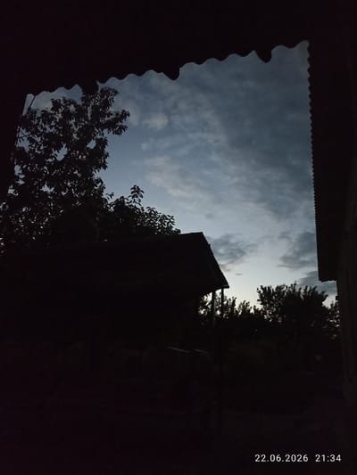 The image depicts a silhouetted view of a rural or suburban landscape at dusk. In the foreground, dark, silhouetted structures, possibly a roofline and parts of a building, frame the scene from the top and right. A prominent dark silhouette of a house with a sloped roof is visible in the mid-ground, with trees behind it. To the left, a large tree with dense foliage is silhouetted against the sky. The sky itself is a dusky blue with scattered clouds, suggesting the transition from day to night. Trees line the horizon in the mid-ground and further into the background. A timestamp in the lower right corner reads "22.06.2026 21:34", indicating the date and time the photograph was taken. The image appears to be taken from a sheltered position, looking out over an outdoor area. No people or specific activities are visible. The environment is dark and shadowed, with the sky being the primary source of light. This scene is consistent with a location in Batken City, Kyrgyzstan, given its rural or semi-rural characteristics and the timestamp.