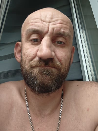 A close-up selfie of a bald, middle-aged Caucasian male with a beard and wearing a silver chain is taken from the chest up. The man is looking directly at the camera with a neutral expression. His skin is fair with some texture and small blemishes. The background shows a window with blinds and a view of a building. The lighting suggests it is daytime. The location is Stargard, Poland.