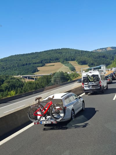 This media file depicts a scene on a highway in Germany, likely during daytime given the bright sunlight and clear blue sky. The primary subjects are vehicles stopped in traffic. In the foreground, a white car is parked, and a bright red bicycle is mounted on a rack at the rear of the car. The license plate of this car is visible and reads "KS PR 293". Further ahead, a white van is also stopped, carrying a yellow bicycle on its rear rack. Several other vehicles are visible in the background, indicating a traffic jam.
The setting is a highway with lush green forested hills on the left side and a clear blue sky above. A large bridge or viaduct is visible in the distance, spanning across a valley. The terrain suggests a rural or mountainous region. The weather appears to be sunny and warm. The overall mood is one of a typical travel day interrupted by traffic congestion. There are no people clearly visible in the media file. The image is captured from a slightly elevated perspective, looking down onto the highway.