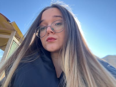 The image is a low-angle, head-and-shoulders selfie of a female. She possesses long, straight hair with prominent blonde highlights, strongly backlit on the right, creating a bright halo effect. Her facial features include light-colored eyes behind round, thin-framed eyeglasses, and lips colored with a reddish-pink tint. She is wearing dark-colored attire, possibly a jacket. Her gaze is directed straight at the camera with a neutral expression. The background features a clear, bright blue sky dominating the upper right. In the upper left, a segment of a yellow-painted structure, possibly an overhang or eave with visible support beams, is present, alongside a faint glimpse of a light green wall section below it. The lighting indicates an outdoor daytime setting under direct sunlight. The specific location within Bol'shevik, Russia, is not identifiable from the visual information contained within the image.