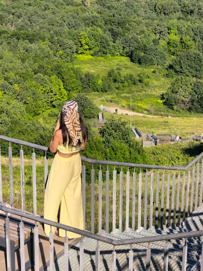 A woman, viewed from behind, stands on a metal staircase overlooking a lush, green hillside covered in trees and grass. She wears a yellow top and wide-leg pants, accessorized with a striped headscarf. The background shows a vast expanse of dense forest that transitions into a grassy area with scattered wooden structures and pathways. The image is taken during the daytime, with sunlight casting shadows on the staircase. The location is identified as Akhmerovo, Russia.