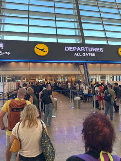 The image displays a detailed view of a busy departures area within an airport terminal, specifically identified as Ben Gurion Airport (TLV), Israel, based on the visible trilingual signage. The scene is illuminated by natural light filtering through a high, grid-patterned glass ceiling, complemented by artificial overhead lighting and a prominent blue LED strip light beneath the main sign.
The central overhead dark gray sign clearly indicates "DEPARTURES ALL GATES" in white lettering, flanked by yellow circular logos each featuring a black airplane silhouette. Arabic text is also visible on the left side of the sign.
Approximately 30-40 individuals, primarily adults, are visible, queued in multiple lines defined by silver stanchions and blue retractable belts on a light-colored tiled floor. Most individuals are facing away from the camera, progressing towards the background. They are casually dressed in various attire, including t-shirts, shorts, and jeans, and are carrying personal belongings such as backpacks and tote bags.
In the immediate foreground, the back of a person with reddish-brown hair is visible, wearing a dark top with purple and yellow accents. Directly in front of them, a blonde-haired individual wears a white top and carries a floral-patterned tote bag and a light brown shoulder bag. To their left, a male in a yellow t-shirt and shorts wears a large orange-brown backpack.
Further into the background, multiple security screening stations or check-in counters are discernible, equipped with dark-colored conveyor belts and other processing equipment. Several cylindrical metallic trash receptacles are positioned alongside the queue lines. The overall scene depicts an active passenger flow typical of a major airport's departure operations.