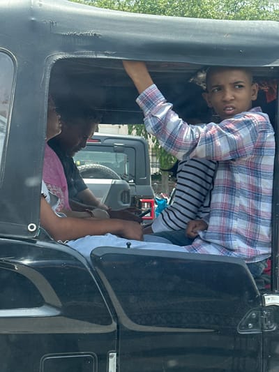 The image captures a scene inside a black auto-rickshaw in Ajmer, India. Three people are visible. In the foreground, a young boy with a shaved head is looking to the right, his mouth slightly open, and he is wearing a plaid shirt. To his left, an adult male with dark skin and short black hair is looking down, wearing a dark t-shirt. Between them, a person wearing a pink garment with a patterned hem is partially visible. The interior of the auto-rickshaw appears worn, with visible damage to the fabric roof. Outside the vehicle, a glimpse of trees and another vehicle, possibly a grey SUV, can be seen. The overall lighting suggests a bright, sunny day.