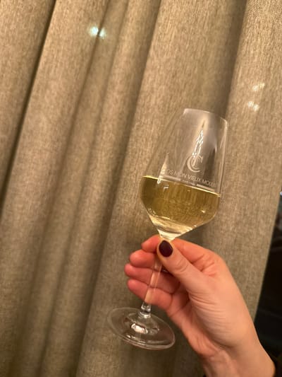 A close-up shot features a hand holding a wine glass against a textured fabric background. The hand, appearing to be female, has dark plum or burgundy nail polish on the thumb and visible fingers, and holds the clear glass by its stem. The glass is about half-filled with a light, pale yellow liquid, consistent with white wine or champagne, showing various reflections from the ambient lighting.
The background consists of a soft, woven fabric, possibly drapes or upholstery, in a warm, muted beige-brown color, characterized by prominent horizontal folds and subtle texture. The lighting is soft and warm, suggesting an indoor setting, possibly in the evening or a dimly lit room.
Visible text on the side of the wine glass reads "CLOS MON VIEUX MOULIN" with "CUVEÉ PARIS" below it. A stylized logo, potentially two interlocking 'C's or 'J's, is positioned above this text. The image is oriented such that the glass and hand are positioned horizontally across the frame, though the object itself is likely held upright. While the provided context mentions "Grass, Luxembourg," the image itself shows an indoor scene with no visible outdoor elements.