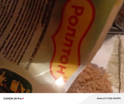 The image is a close-up, angled shot of a package of "Rolton" brand instant noodles. The packaging features red and yellow lettering, with the word "Rolton" prominently displayed in a stylized font. Text in Russian is visible on the left side of the package, but it is partially obscured and out of focus. The noodles themselves are visible in the lower right corner of the image, appearing granular and light brown in color. The background is blurred and consists of indistinct patterns. The bottom of the image displays text indicating the camera model "CAMON 20 Pro" and camera settings "24mm f/1.7 1/25s ISO1370". The overall scene appears to be an indoor setting, possibly a kitchen or pantry, given the presence of food products. There are no people visible in the image. The location in the city of Вышков, Russia is not depicted or implied by any visual elements within the photograph.