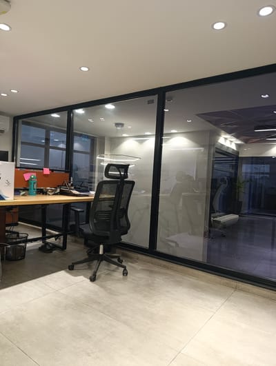 The image depicts an office interior, specifically a desk area with a glass partition wall. The desk is made of light-colored wood and has a black office chair with wheels in front of it. On the desk, there is a black document holder, a light blue water bottle, and some sticky notes. To the left of the desk, there is a trash can and a box. The floor is tiled with large, light-colored square tiles. The office appears to be modern, with recessed ceiling lights. The glass partition offers a view into another section of the office, where more desks and chairs are visible, along with decorative lighting fixtures. The image was taken in Lidher, Pakistan, based on the provided information.