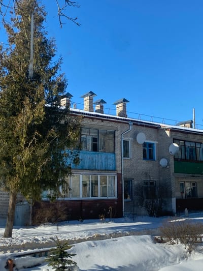 The image depicts a multi-story residential building in Карабоўскі сельскі Савет, Belarus, under a clear, cloudless blue sky during winter. The foreground and ground surrounding the building are covered in white snow. A tall, dark green coniferous tree, displaying visible icicles, dominates the left side, partially obscuring the building's facade.
The building is constructed of light-colored brick and presents at least two visible floors. Its roof carries a thin layer of snow and features four brick chimneys with dark, pyramidal caps, in addition to a single metal pipe further to the right. A white vertical drainpipe is visible on the facade.
On the upper floor, an open balcony with a distressed, light-blue wooden railing is positioned next to an enclosed loggia. The ground floor includes a prominent enclosed loggia with white-framed windows and a dark brown lower panel. Various windows with white and blue frames are scattered across the facade, including a distinct blue-framed window on the upper right. Three white satellite dishes are mounted on the building's exterior. A small, light-colored utility door is located on the ground level, to the left of the large tree. A small coniferous sapling is partially visible in the snow in the lower foreground. No people or active interactions are observed within the scene.