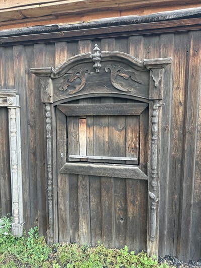 This is a medium shot, eye-level photograph of a weathered wooden structure in the city of Nogornyi, Russia. The central subject is a closed wooden door or window with a decorative wooden frame. The frame is elaborately carved with what appear to be two stylized birds facing each other above a rectangular opening. The wood is dark and aged, showing signs of significant wear and tear, with visible grain patterns and some cracks. There is a horizontal wooden plank positioned across the opening of the central frame. Flanking the central frame are two vertical decorative wooden elements, one to the left and one to the right, both with carved, bead-like segments. To the far left, a portion of another decorative wooden element, possibly a pillar or post, is visible, appearing lighter in color and also showing signs of age. The backdrop is a wall constructed of vertical wooden planks, also weathered and dark. At the bottom of the image, green vegetation, likely grass and small plants, is growing, with some dark debris, possibly coal or ash, mixed in with the soil. The lighting suggests it is daytime, but the sky is not visible.
