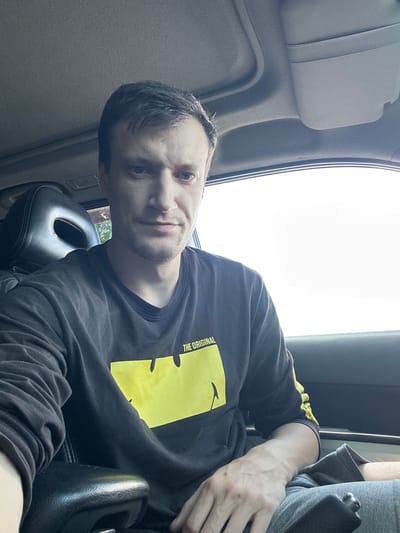 A young man is seated in the passenger seat of a vehicle, with the camera angled from the front passenger side, looking towards the driver's side. He is wearing a black long-sleeved shirt with a prominent yellow graphic on the chest that reads "THE ORIGINAL". The man has short, dark hair and is looking directly at the camera with a slight smile. The interior of the car is visible, including the headrest and part of the dashboard. The lighting suggests it is daytime, possibly late afternoon, with bright light coming from outside the vehicle, creating a slight glare. The image appears to have been taken in Shatava, Ukraine.