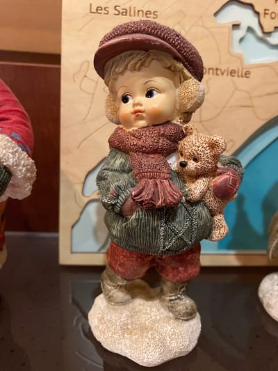 This image features a figurine of a child holding two teddy bears. The figurine is light-skinned with rosy cheeks and blue eyes, wearing a burgundy hat and a green coat with red accents. One teddy bear is brown and the other is also brown with darker brown stitching. The figurine is positioned on a wooden surface with a map-like design. Visible text on the map includes "Les Salines" and "montvielle". The background is a dark brown surface. The lighting appears to be indoor, with no discernible indication of time of day or weather. The setting seems to be a decorative display or shelf.