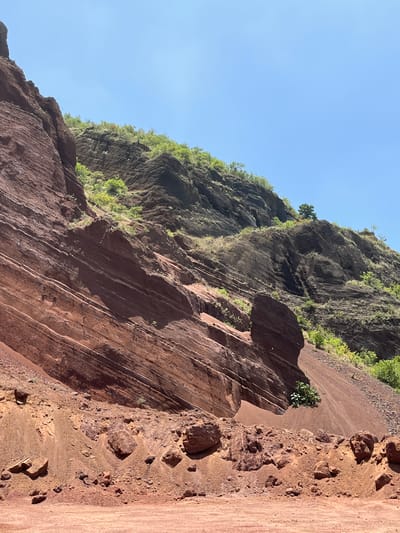 The image depicts a natural landscape of eroded red rock formations and slopes under a clear blue sky. The foreground features loose reddish-brown soil and scattered rocks. Behind this, a steep, layered red rock cliff face dominates the left side of the frame, showing distinct strata. To the right and further back, a darker, more rugged mountainside is visible, covered in patches of green vegetation. A single tree stands atop this darker slope, with another smaller tree visible in the distance on the right. The scene appears to be a natural geological formation, possibly a canyon or hillside, with evidence of erosion and weathering. The location in San Miguel Papaxtla, Mexico, implies a terrestrial environment with arid or semi-arid conditions that would support such rock formations and sparse vegetation. There are no people, animals, or man-made structures clearly visible in the image.