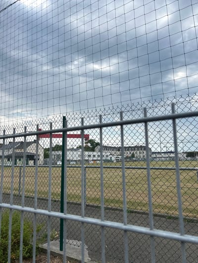The image, taken from behind a chain-link fence, looks out onto a sports field in Saint-Nazaire, France. The field itself is predominantly brown, suggesting dry grass, and is bordered by a paved path. In the background, several modern white buildings with dark roofs are visible. A prominent red crane, likely associated with construction, stands out against the cloudy sky. The foreground is dominated by the silver bars of the chain-link fence and a black net that stretches across the frame, obscuring parts of the scene. A green metal post from the fence is also visible. The overall environment suggests an outdoor sports facility or recreational area within an urban setting, under a cloudy, overcast sky.